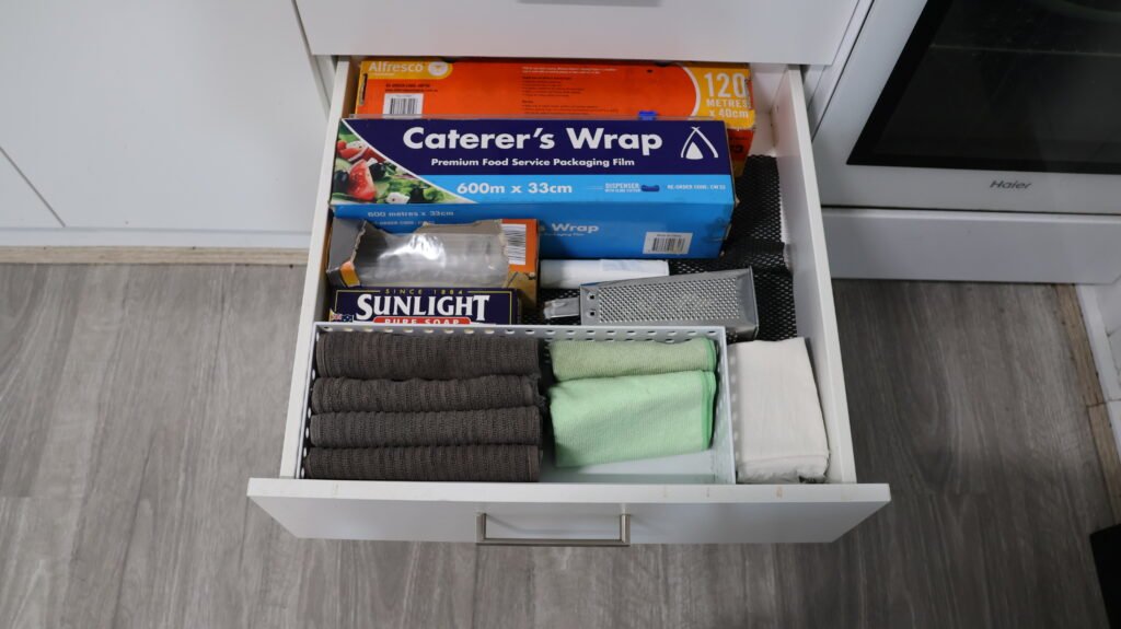 An open kitchen drawer holding five folded grey tea towels, a few cleaning cloths and kitchen wraps — slow decluttering cut to what one day of washing needs