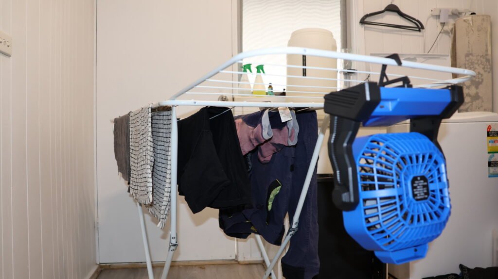 Clothes drying indoors on a folding airer with a blue fan clipped to it